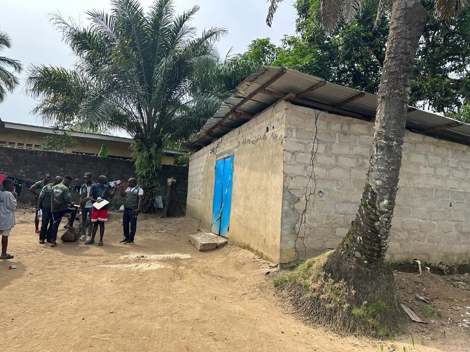 Environmental Protection Agency inspectors talk with the operator of C3M water factory outside the closed building, identifiable by its blue gate and unpainted exterior.